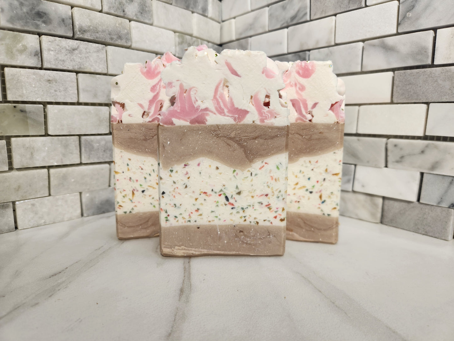 Three layered handmade soap bars with pink floral tops and multicolor sprinkles, displayed on a marble surface against a gray brick wall.