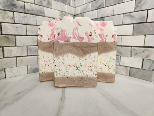 Three layered handmade soap bars with pink floral tops and multicolor sprinkles, displayed on a marble surface against a gray brick wall.